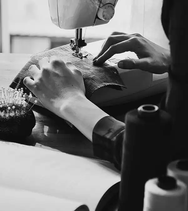 Close das mãos de uma costureira operando uma máquina de costura, com alfinetes e linhas na composição. Imagem em preto e branco.